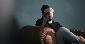 Foto que muestra a una persona sentada sobre un sofá con una mano sobre su cara y una expresión de tristeza y agobio